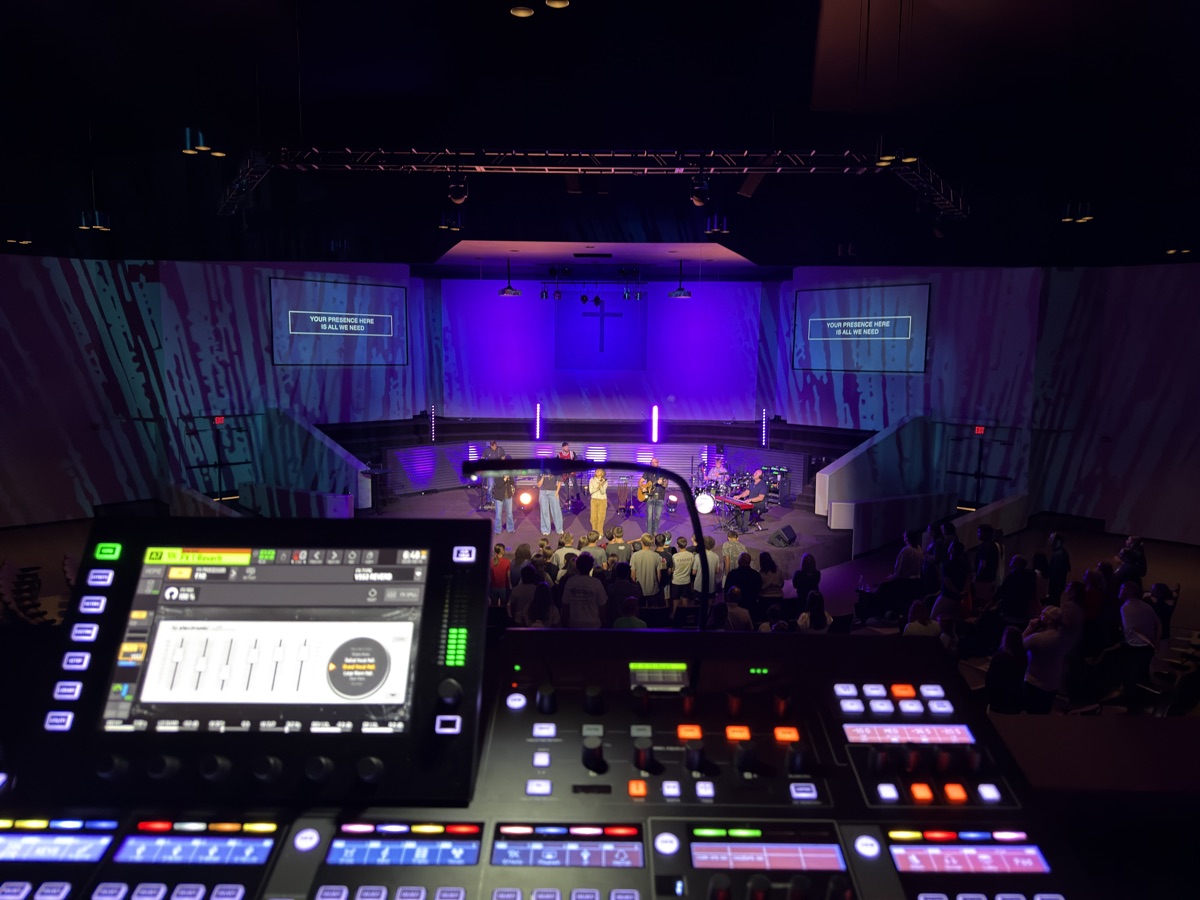 Mixing console overlooking a worship band on a stage lit in purple